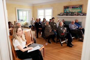 A group of people attend a meeting or presentation in a residential living room with hardwood floors and beige walls.