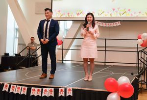 Two speakers stand on a stage decorated with red and white balloons and banners for a celebration event.
