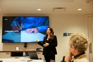 A presenter in a black blazer gestures while speaking during a business presentation in a conference room.