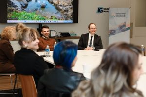 Business meeting participants seated at a conference table discussing while a nature documentary plays on screen.