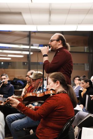 A musician in a burgundy sweater performs with microphone while string players in red shirts rehearse in an indoor space.