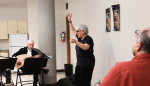 A person gestures while leading a small group session in a plain room with another person sitting at a keyboard.