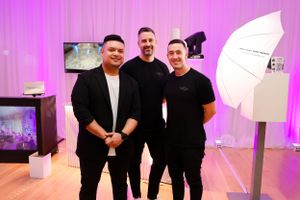 Three people in black attire stand together smiling in a pink-lit studio space with photography equipment visible.