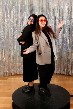 Two people pose playfully in front of a silver tinsel backdrop, one wearing star-shaped glasses and a beige blazer.