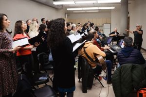 A choir group stands and sings together in a rehearsal room with fluorescent lighting and chairs.