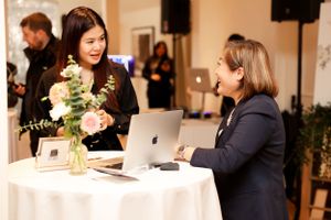 Two people having a professional conversation at a round table with a laptop and floral arrangement at a business event.