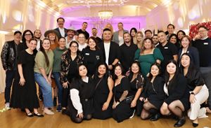 A large group of formally dressed people pose together for a celebratory photo in an elegantly lit ballroom setting.