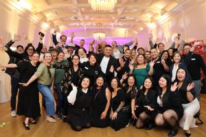 A large group poses energetically together at an elegant event venue with warm lighting and decorative chandeliers.