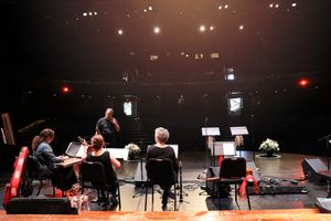Musicians on stage perform in front of music stands during a concert with dramatic stage lighting in the background.