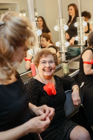 A person wearing a black dress with a red accent sits in front of a dressing room mirror, smiling warmly.