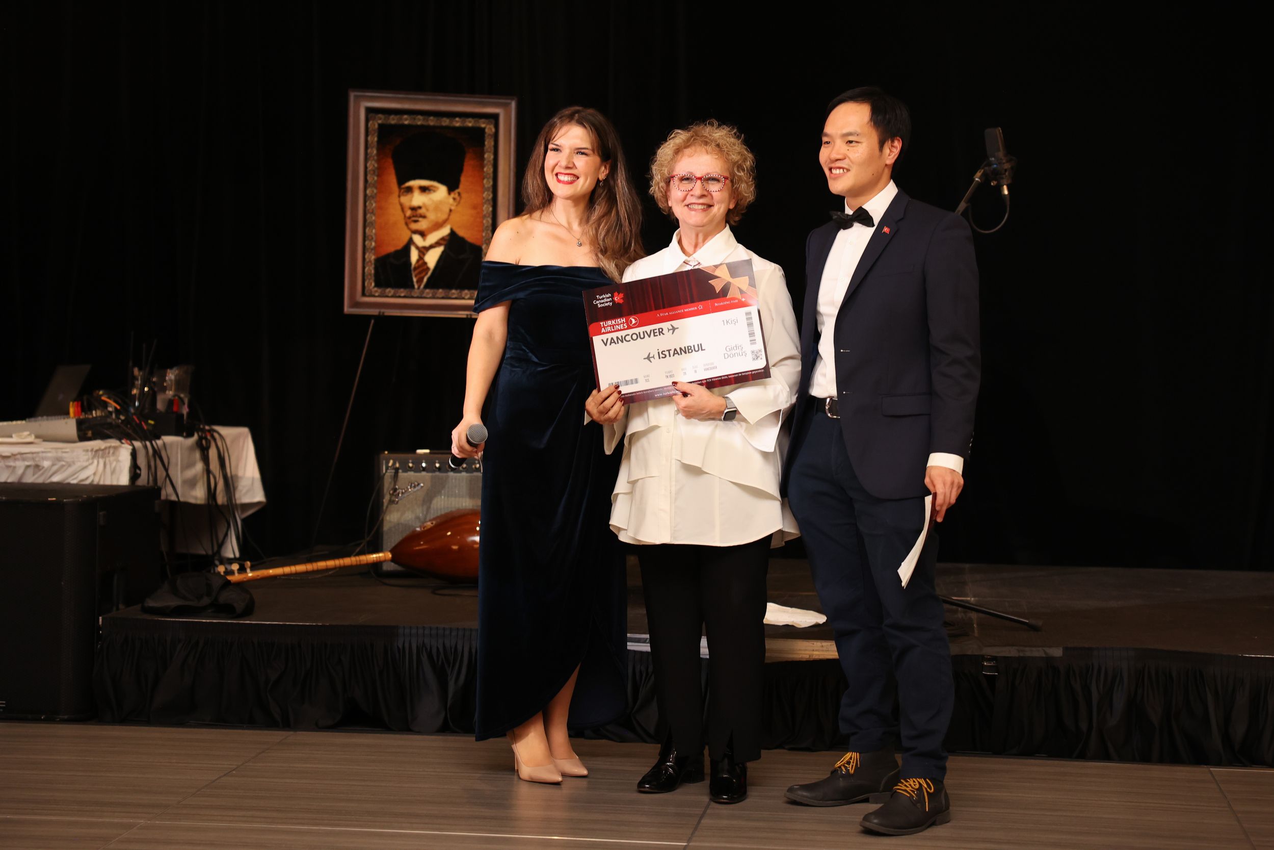 Three people in formal attire stand together on a stage with a portrait painting visible in the background.