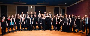 A large choir group in formal black attire stands posed together for a group photo in a performance hall.