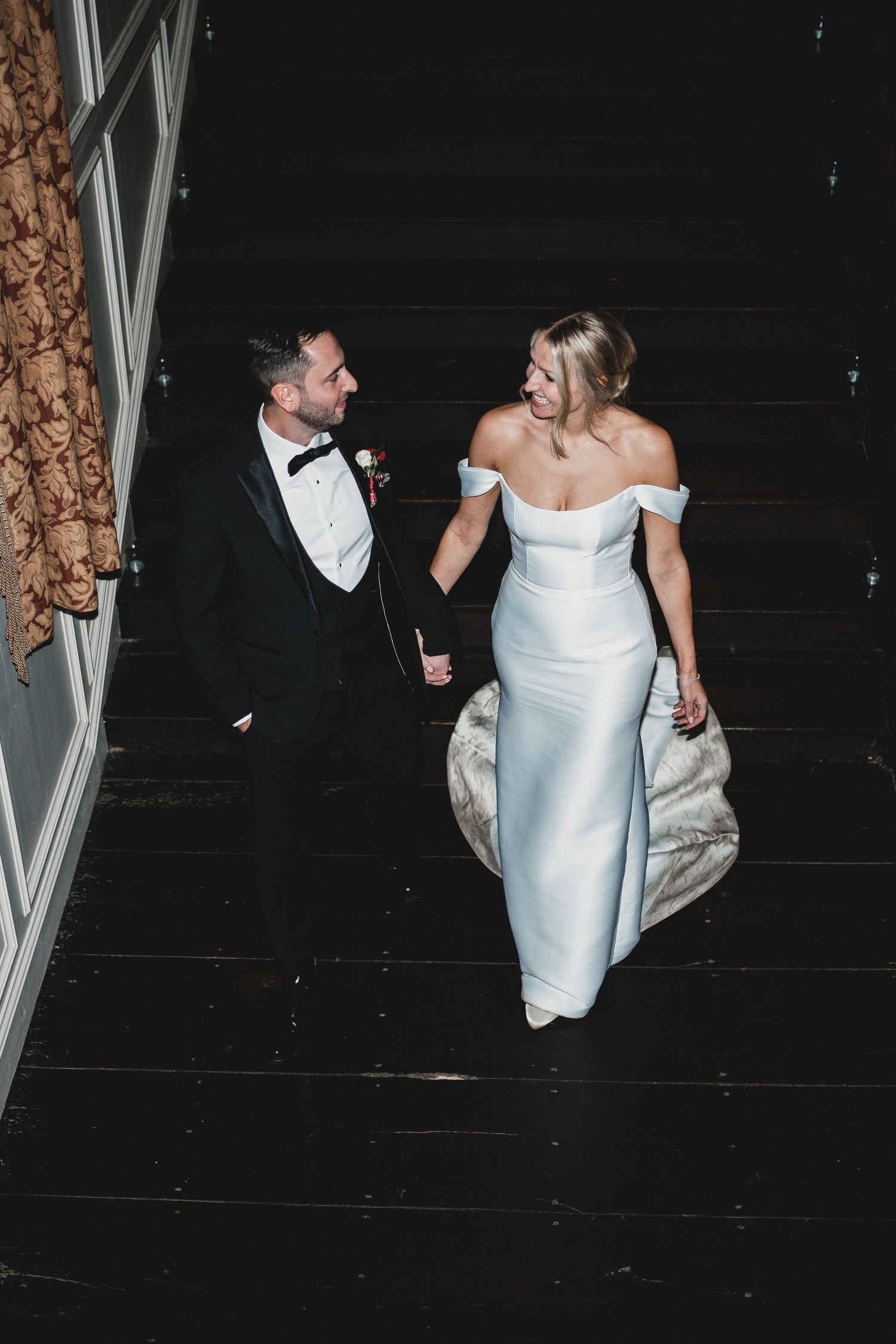 A bride in a white off-shoulder gown and groom in a tuxedo walk together on dark flooring during their wedding celebration.
