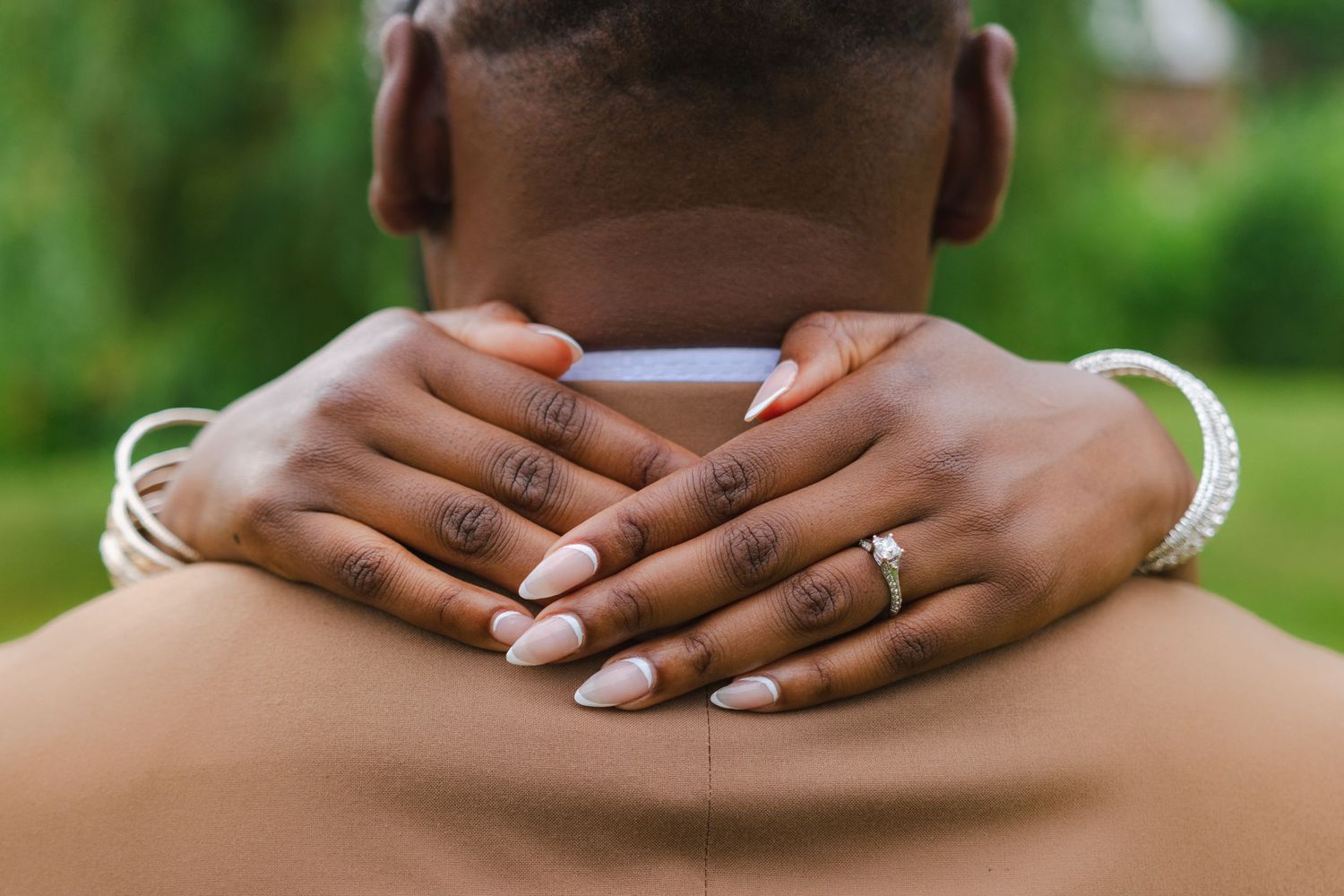 Two hands wearing rings and nail polish embrace around someone's neck outdoors with green foliage in background.