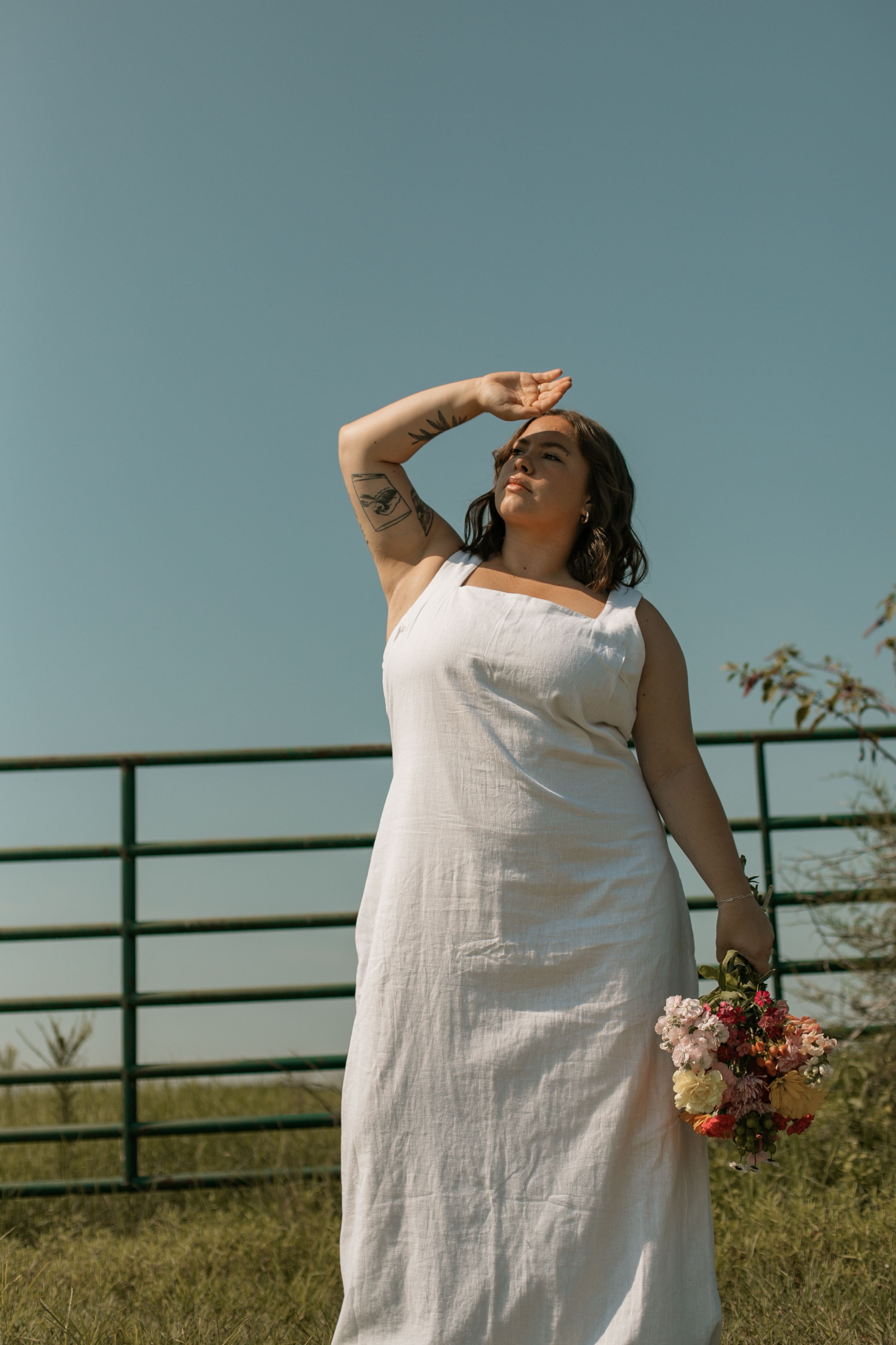 A person in a flowing white dress stands in a sunny field holding a bouquet of flowers while shielding their eyes.