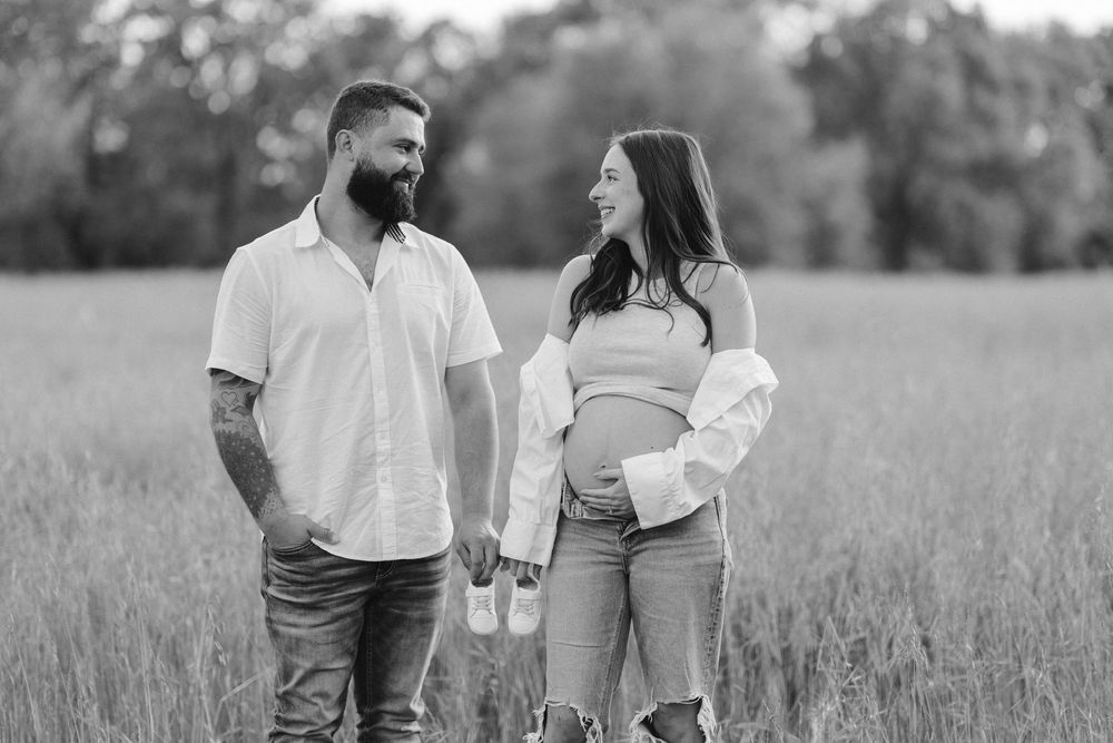A black and white portrait of an expecting couple standing together in a grassy field.