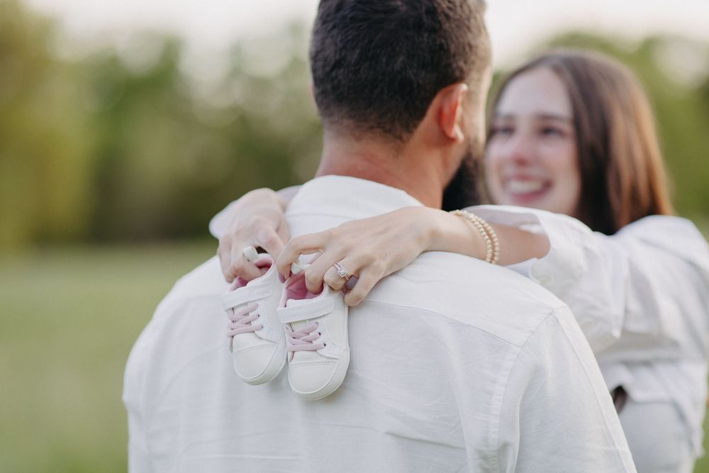 A couple holds tiny baby shoes while embracing in an outdoor setting.