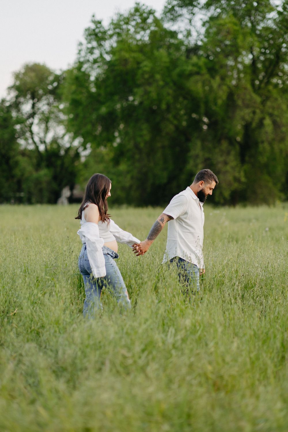 A couple holds hands while walking through a sunlit meadow with lush green trees in the background.