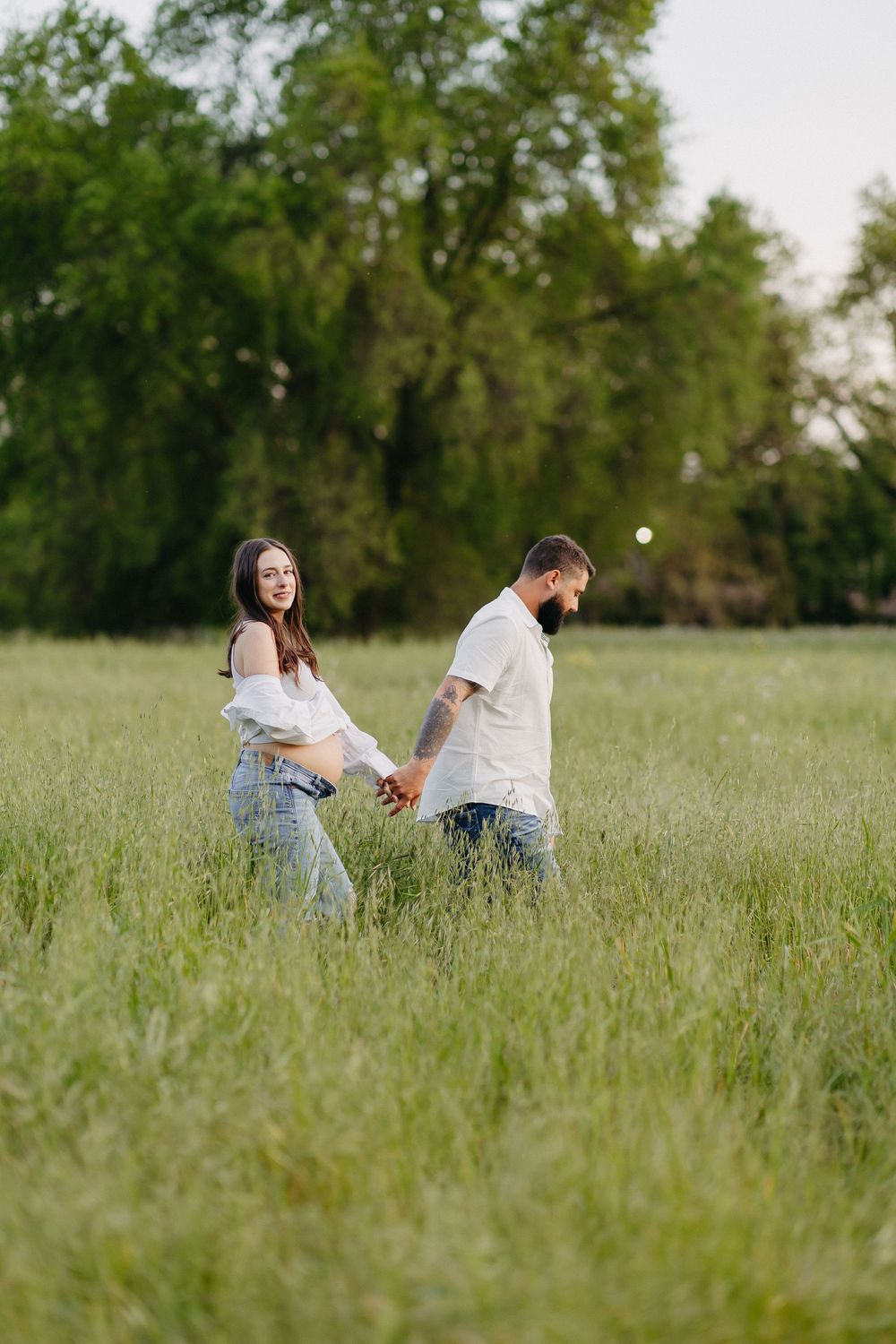 Couple holding hands while walking through a scenic green meadow with trees in the background.