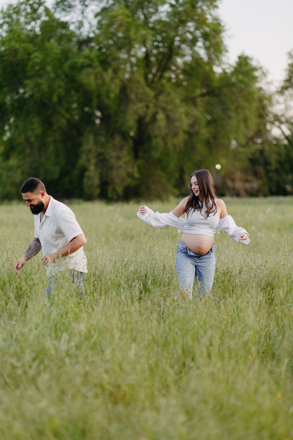 A couple walking playfully through a tall grass meadow with trees in the background on a sunny day.