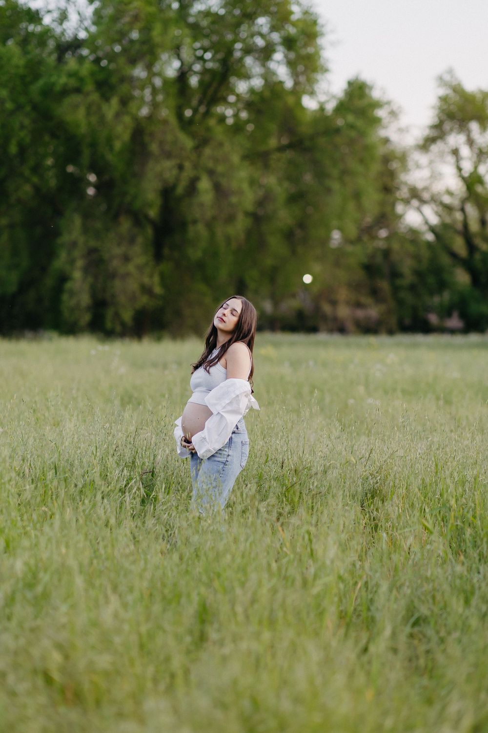 A joyful expectant mother in jeans and white top stands in a sunlit meadow with tall grass and trees.