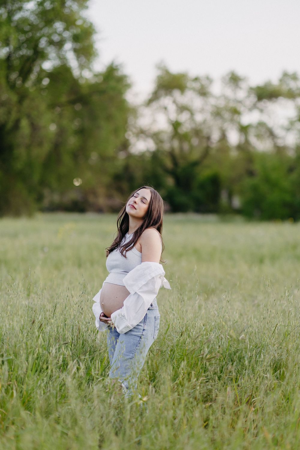 A pregnant woman in casual attire stands joyfully in a sunny field of tall grass.