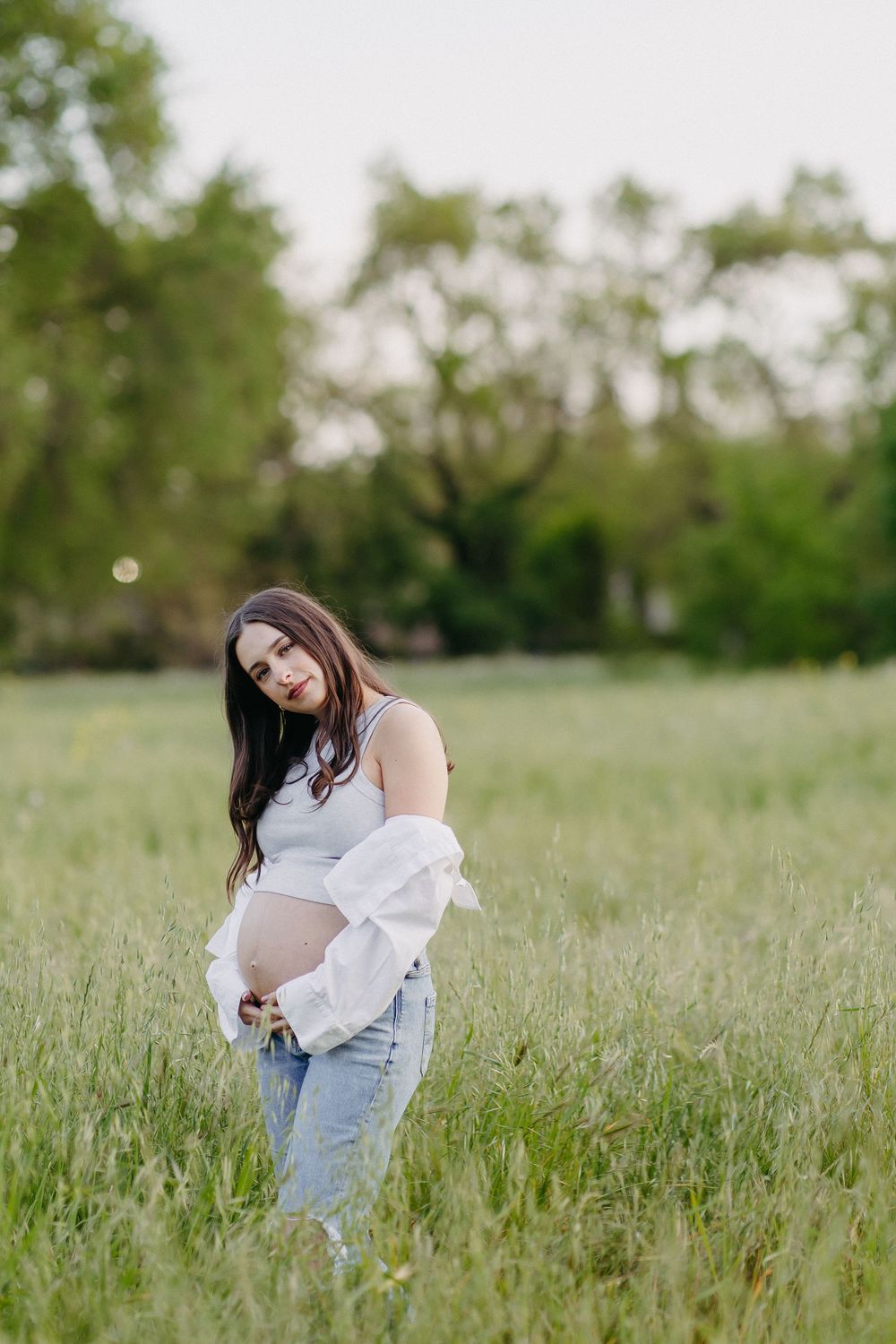 Pregnant woman in casual attire gently holds her belly while standing in a serene field at golden hour.