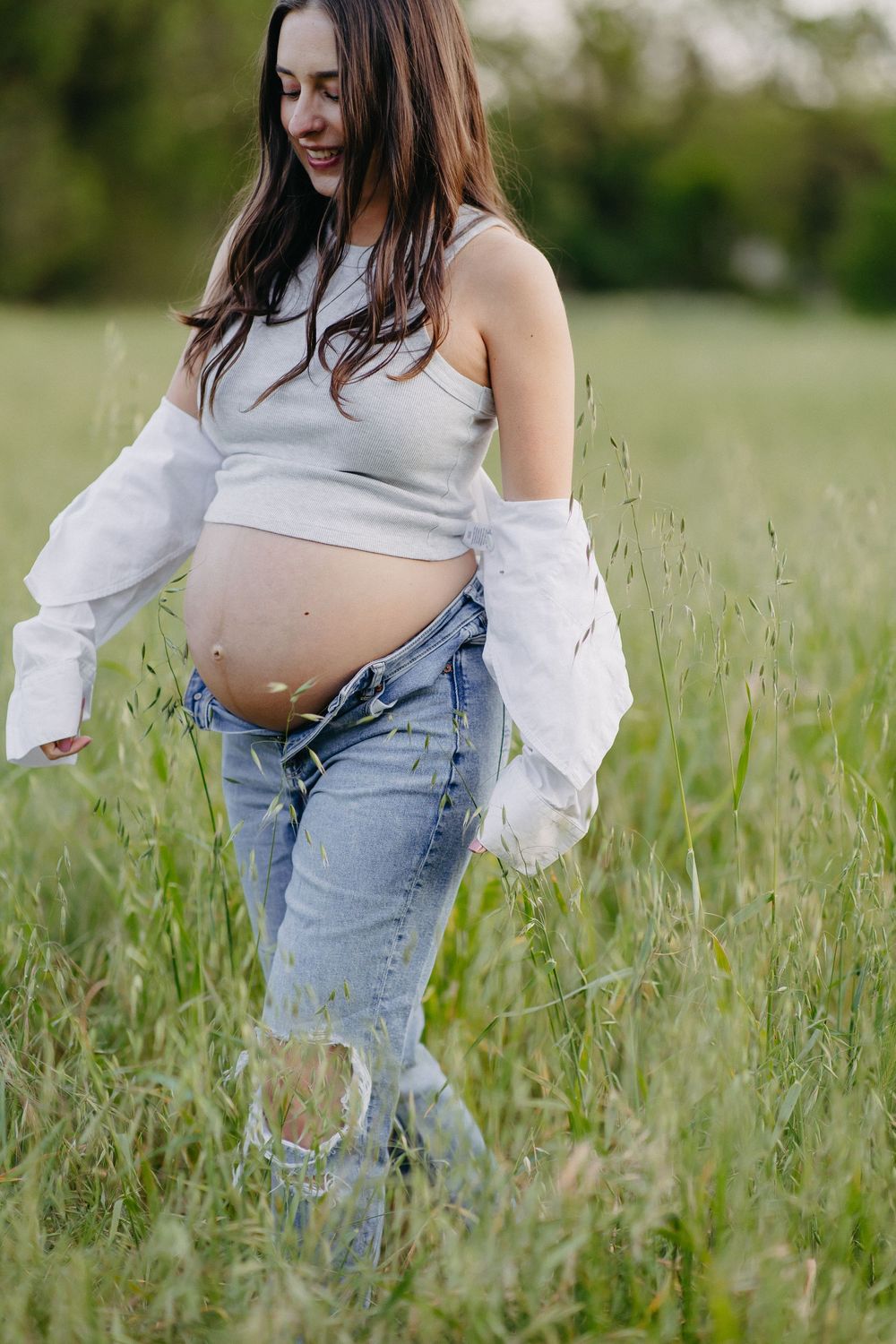 A maternity photo in a field shows an expectant mother wearing ripped jeans and a crop top with flowing sleeves.