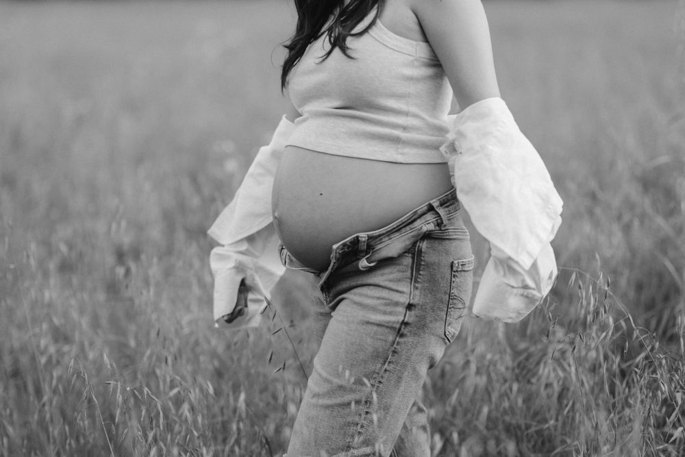Black and white photograph of a pregnant belly in a field with a white flowing shawl and jeans.