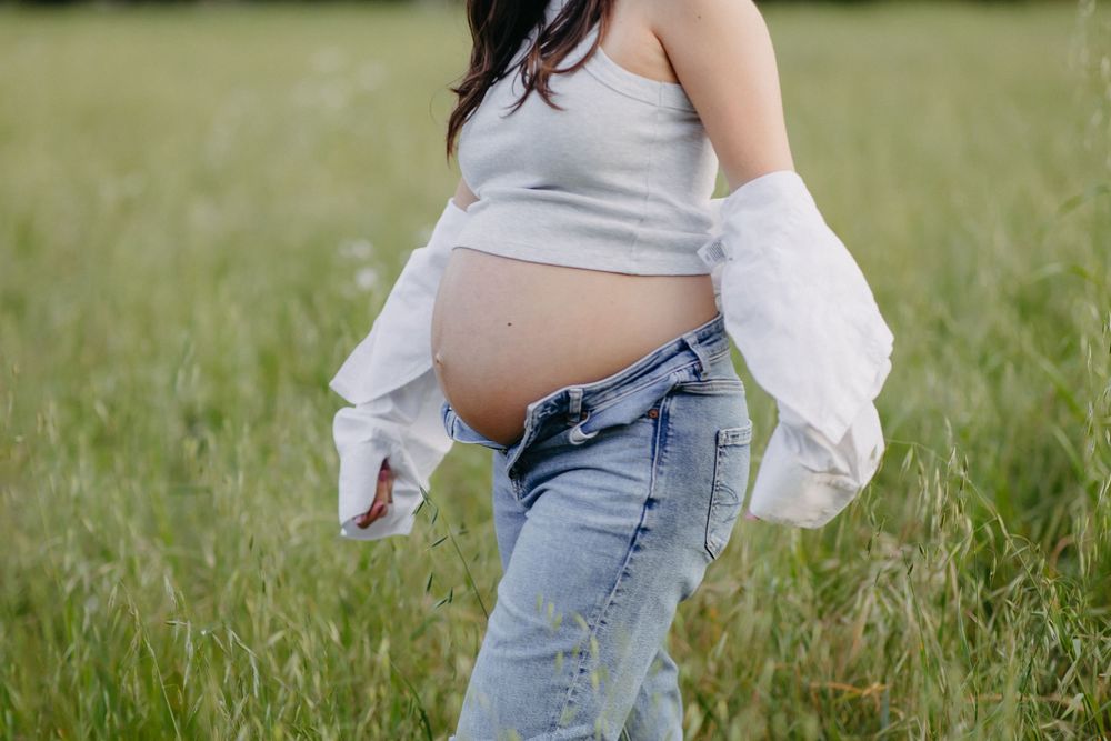 Woman with bare pregnant belly wearing white long sleeve shirt and blue jeans stands in tall grass meadow.