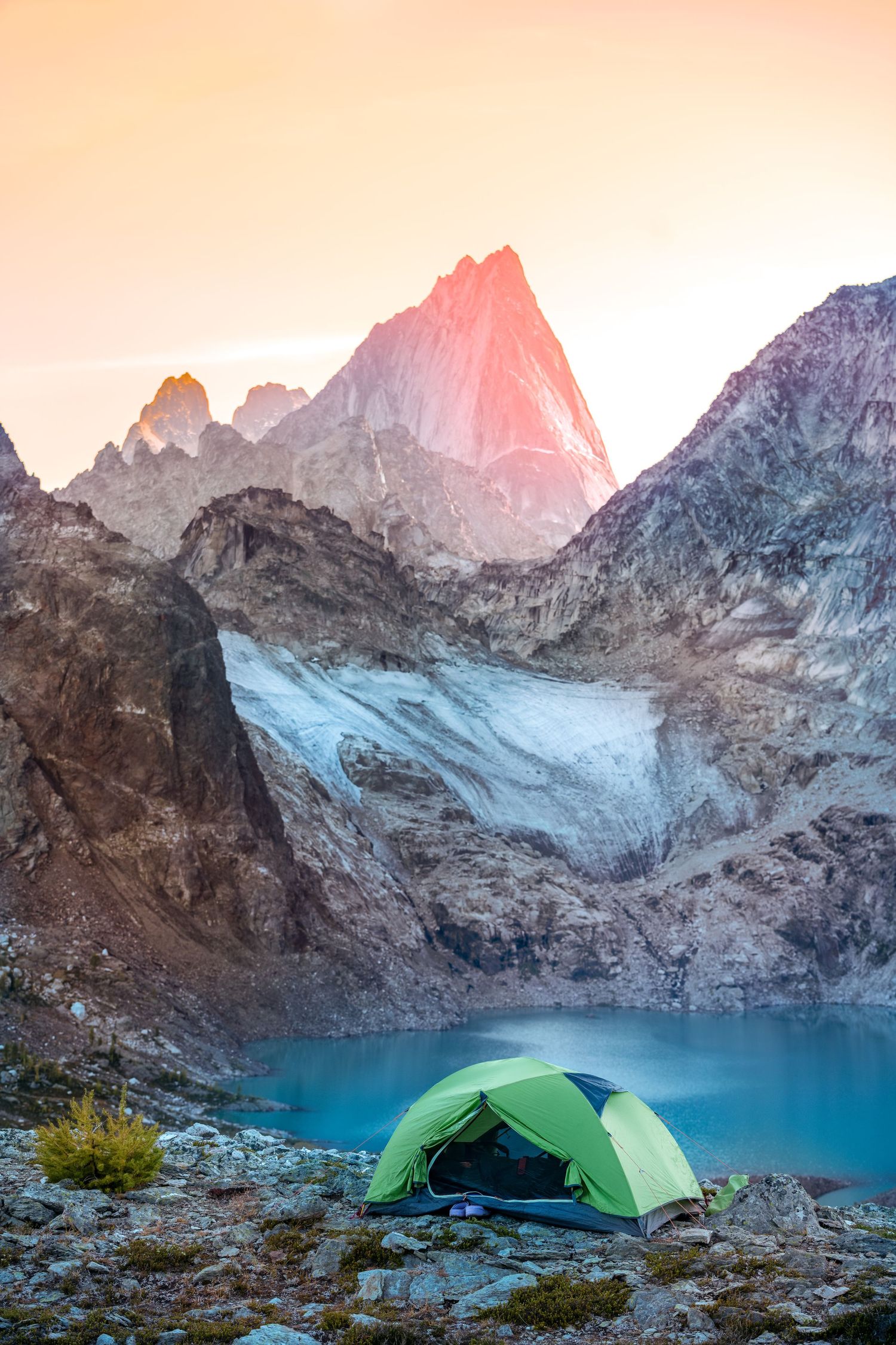 Green camping tent perched near a pristine alpine lake surrounded by jagged mountain peaks at sunset.