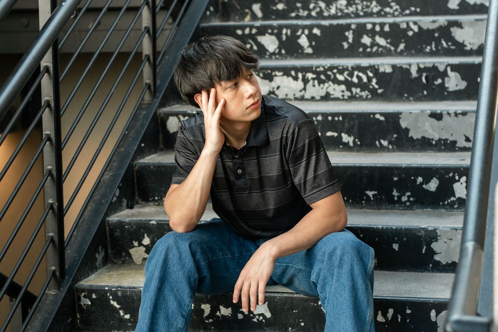 Person sitting on grey steps with head in hand wearing black shirt and blue jeans.