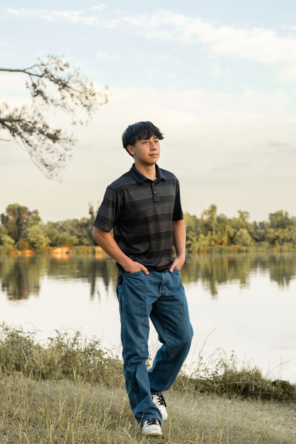 Outdoor portrait by a tranquil lake with trees and greenery in the background, wearing a striped polo and jeans.