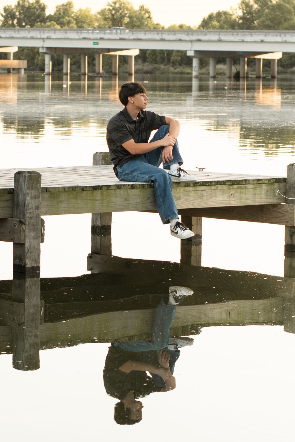 Someone sitting on a wooden dock looks contemplatively across a calm lake at sunset with a bridge in the background.