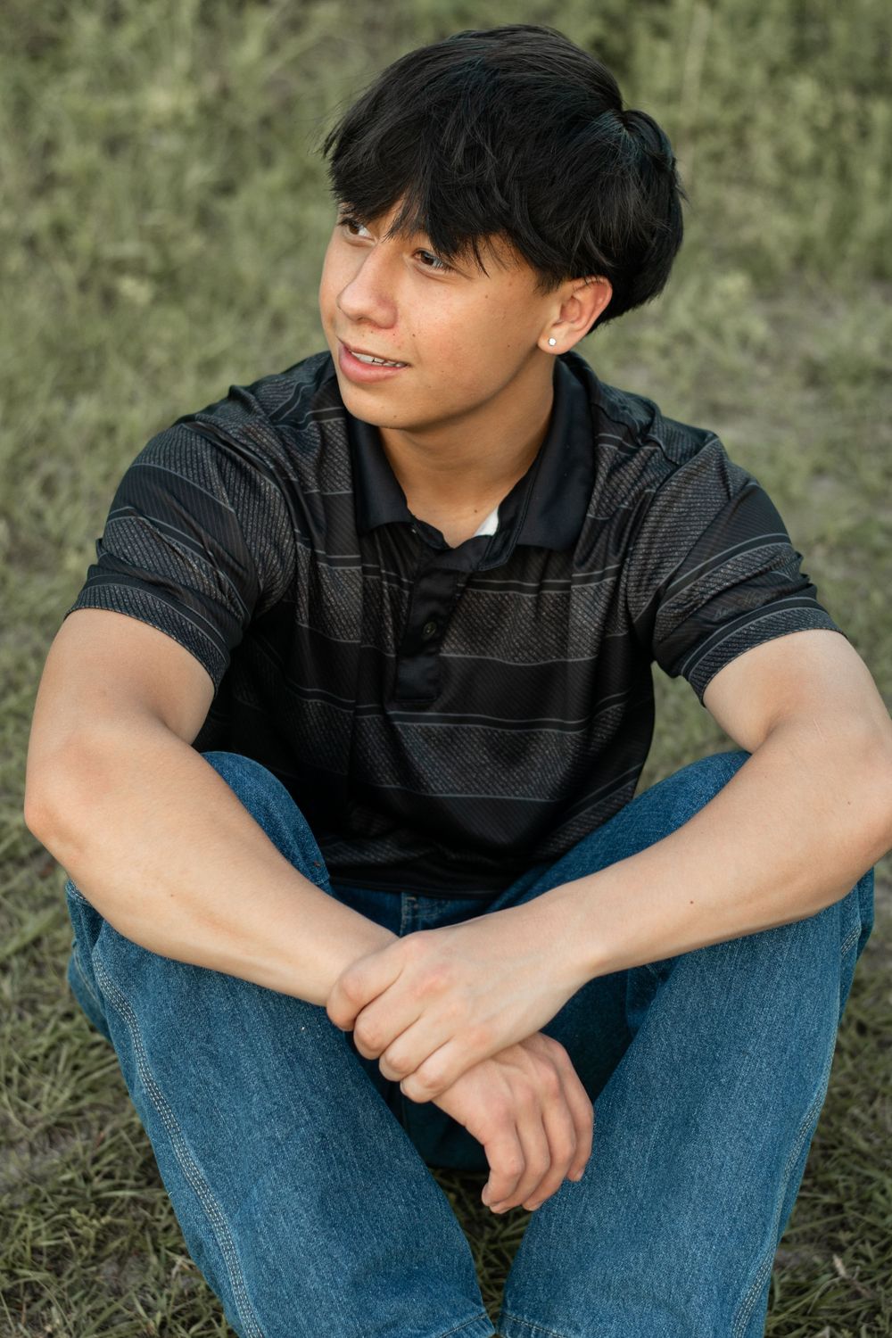 Portrait of person in black striped polo and jeans sitting on grass with a warm smile and relaxed posture.