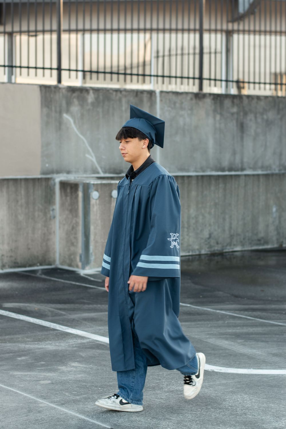 A graduate in a blue cap and gown stands in a parking lot wearing casual sneakers.