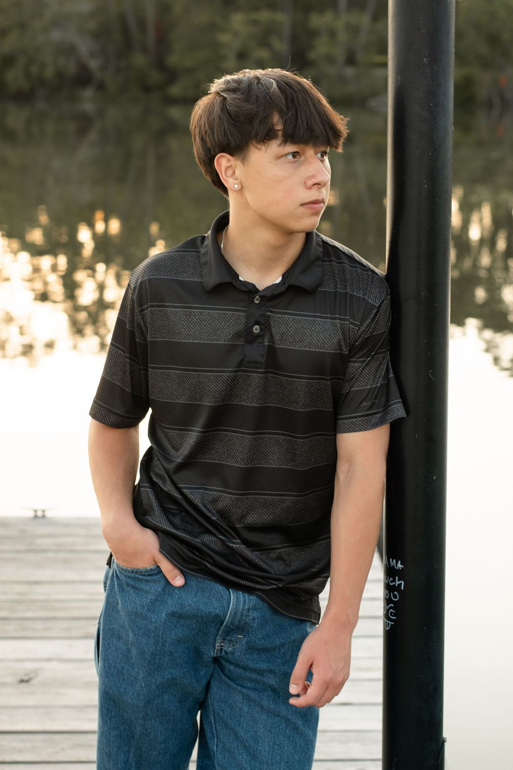 A person in a black striped polo shirt and jeans stands on a wooden dock at sunset.