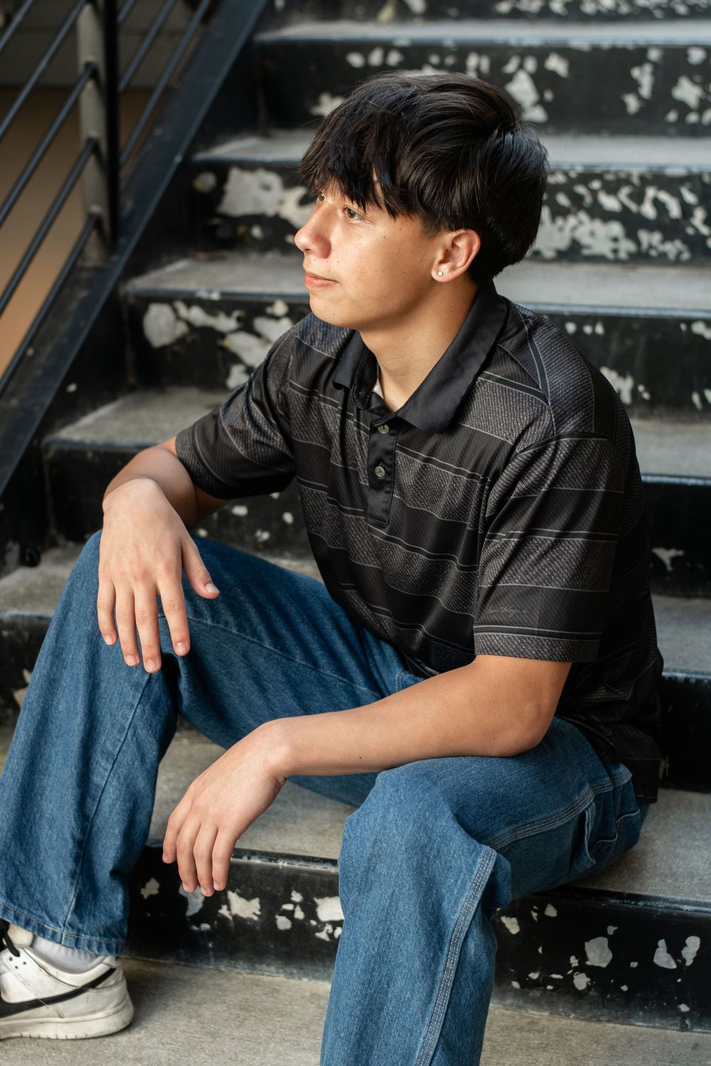 A person in a striped polo shirt and jeans sits thoughtfully on concrete stairs, looking off to the side.