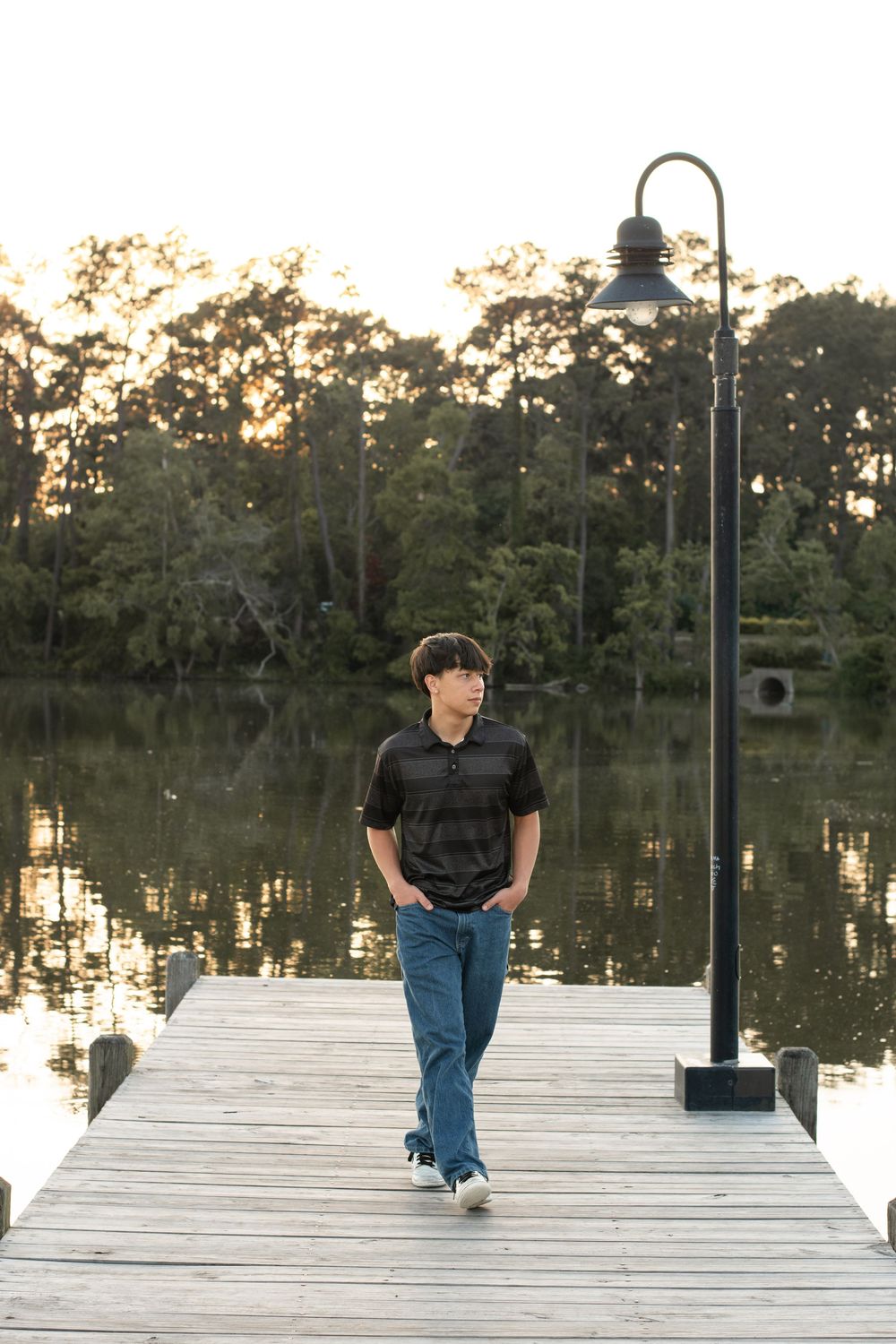 Person walking on wooden dock by lake at sunset with decorative lamp post and trees in background.