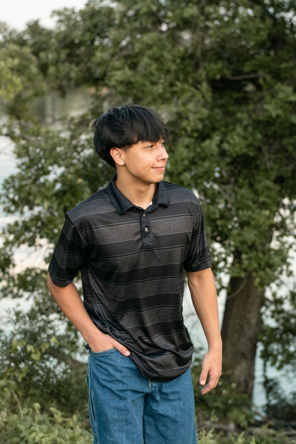 Young person in striped black polo shirt and blue jeans poses outdoors near trees by a lake.