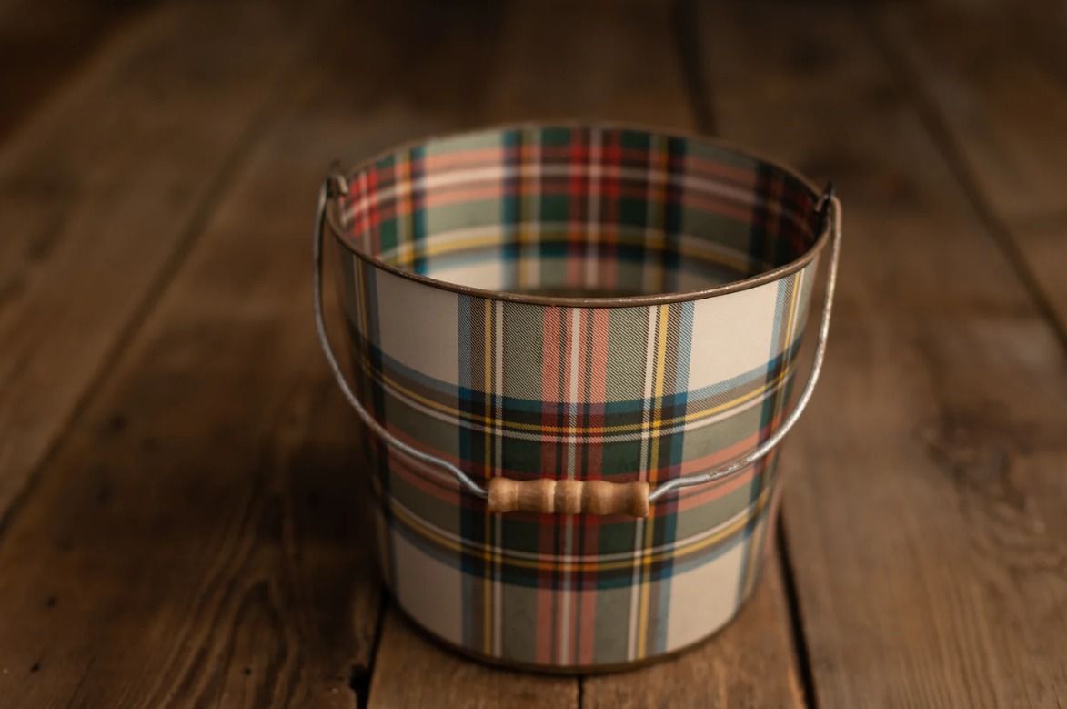 A decorative metal pail featuring a classic tartan plaid pattern with red, green, and cream tones and a wooden handle.