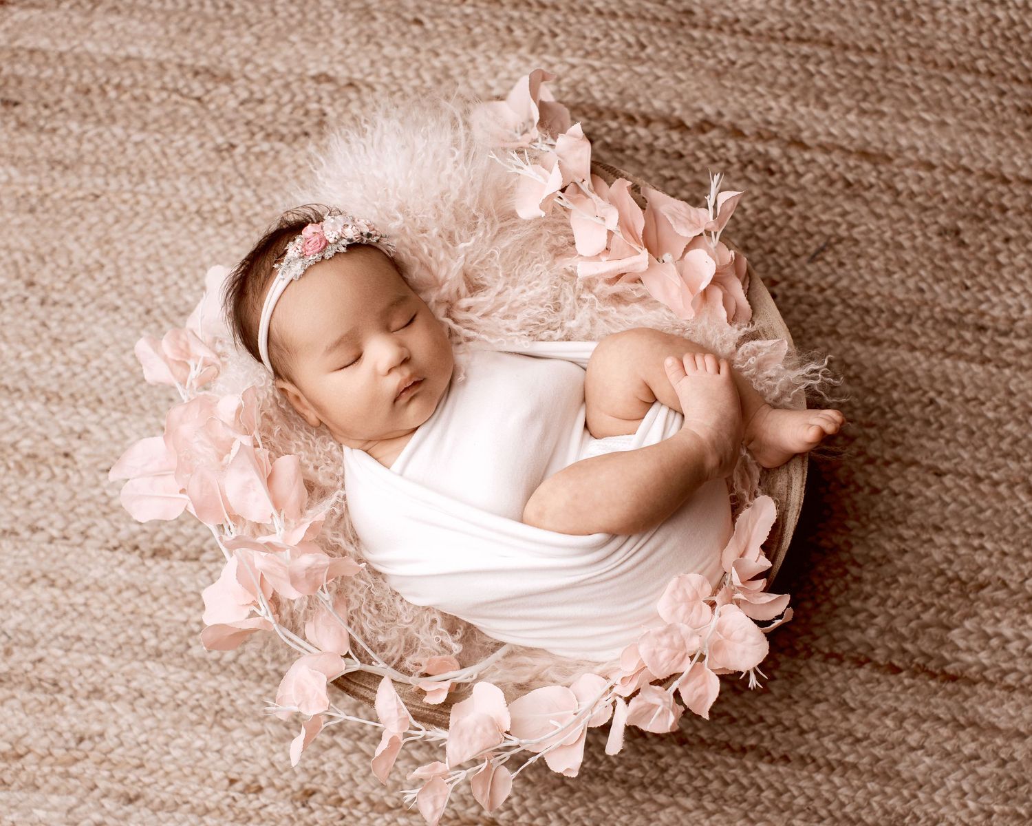 A sleeping newborn baby wrapped in soft white fabric with delicate pink petals arranged around them in a studio setting. Palmerston North