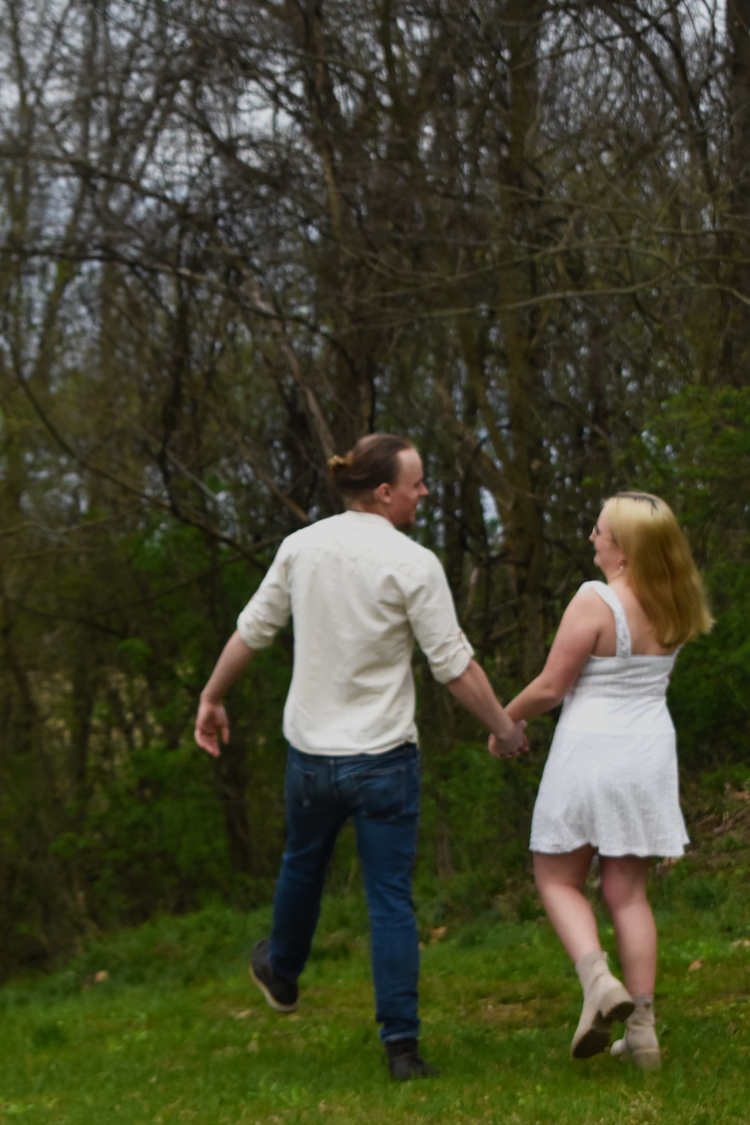 Two people hold hands while walking away on green grass with trees in the background.