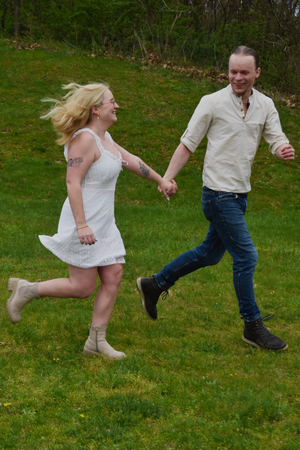 A couple runs joyfully through a green lawn, the woman in a white dress and the man in a light colored shirt and jeans.