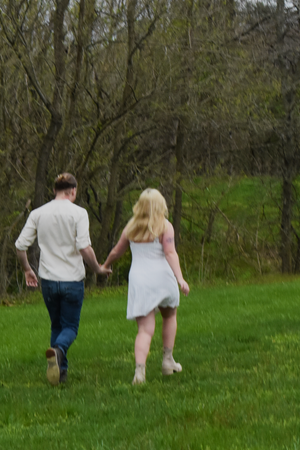 A couple walks hand in hand through a lush green field bordered by trees on a spring day.