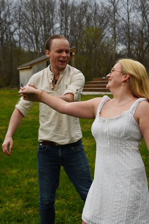 A couple dances and laughs together on a grassy lawn with bare trees and a small shed in the background.