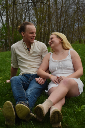 A couple sits together on grass laughing and smiling while wearing casual summer outfits on a spring day.