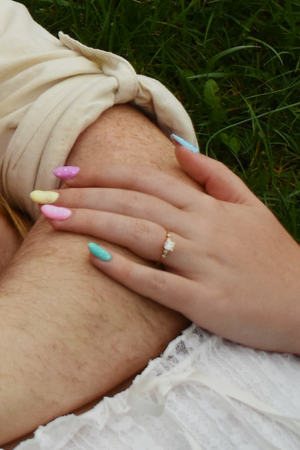 Hand with pastel pink and blue nails wearing a ring rests on beige fabric against green grass background.