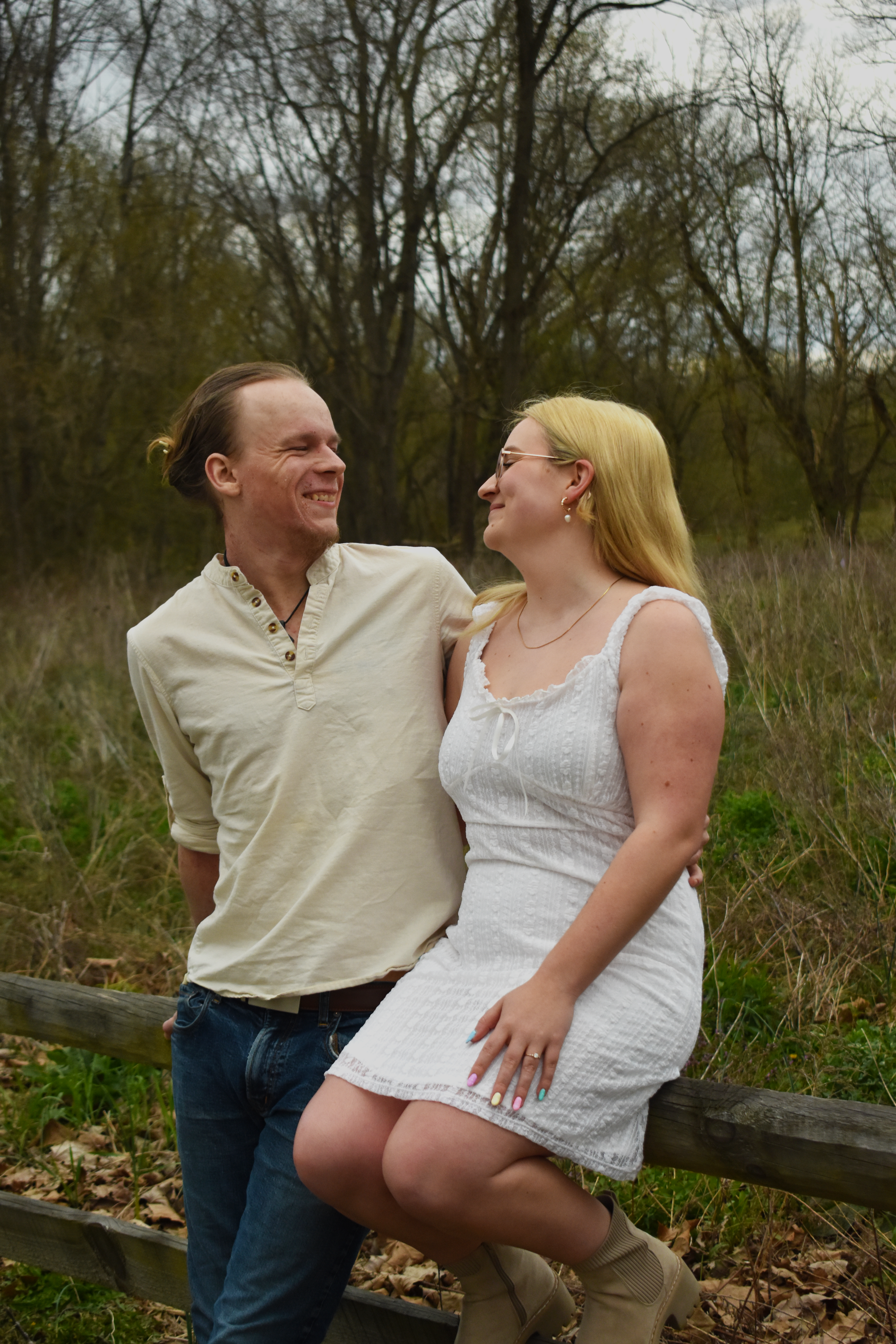 Couple sits on wooden fence in casual attire laughing together during outdoor photoshoot in natural setting.