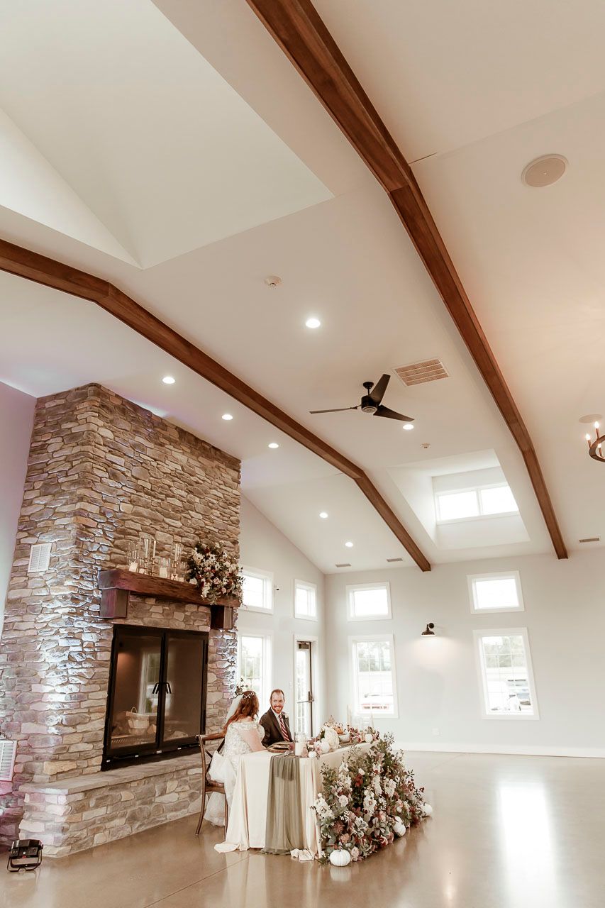 Wedding reception venue with vaulted ceiling, exposed wooden beams, stone fireplace and bright natural lighting.